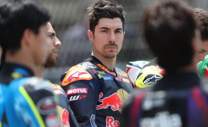 Close-up of a racing rider with dark hair and stubble, wearing a Red Bull KTM racing suit, among teammates.