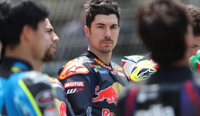 Close-up of a racing rider with dark hair and stubble, wearing a Red Bull KTM racing suit, among teammates.
