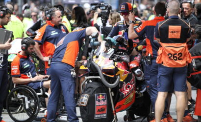 Pit crew in blue-orange uniforms work around a Red Bull KTM racing bike in the pit lane, rider strapped in and teammates assisting various checks and tools nearby.