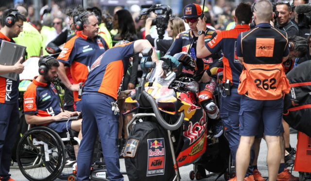 Pit crew in blue-orange uniforms work around a Red Bull KTM racing bike in the pit lane, rider strapped in and teammates assisting various checks and tools nearby.
