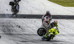 Two riders on a race track; a neon-yellow motorcycle crashes as it leans and lifts its front wheel, debris flying around while another rider watches from behind.