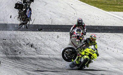 Two riders on a race track; a neon-yellow motorcycle crashes as it leans and lifts its front wheel, debris flying around while another rider watches from behind.