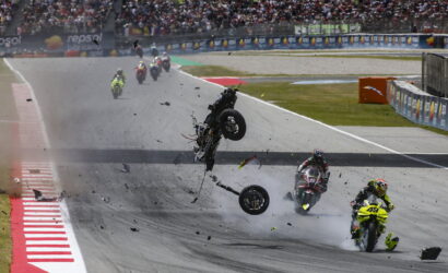 Motorcycle crash on a racetrack with debris flying and riders nearby; spectators watch from the stands.