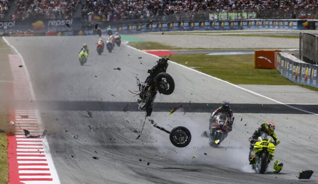 Motorcycle crash on a racetrack with debris flying and riders nearby; spectators watch from the stands.