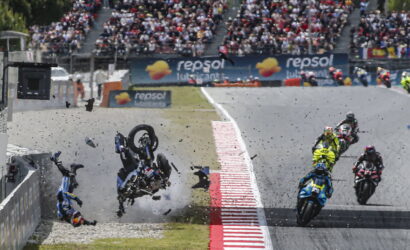 Motorcycle race crash: riders and bikes scattered on track as debris flies, with cheering spectators in stands at the background.