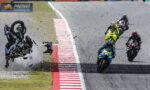 Motorcycle racer flips over at edge of track as others continue racing and debris sprays across the asphalt.