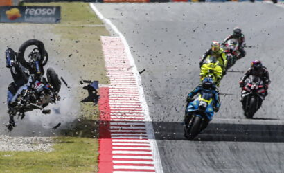 Motorcycle racer flips over at edge of track as others continue racing and debris sprays across the asphalt.