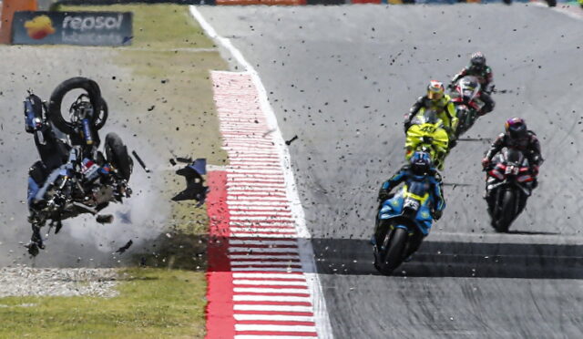 Motorcycle racer flips over at edge of track as others continue racing and debris sprays across the asphalt.