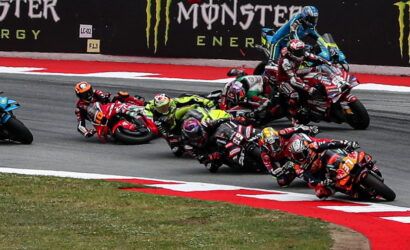 Group of motorcycle racers lean into a sharp turn on a race track, with Monster Energy banners in the background and red-and-white track curbing visible.