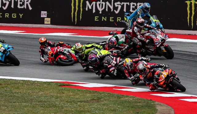 Group of motorcycle racers lean into a sharp turn on a race track, with Monster Energy banners in the background and red-and-white track curbing visible.
