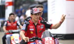 Racing rider in red and black gear on a red bike waves to the crowd, with a teammate on another bike blurred in the background beneath a racing event setting.