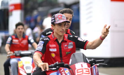 Racing rider in red and black gear on a red bike waves to the crowd, with a teammate on another bike blurred in the background beneath a racing event setting.