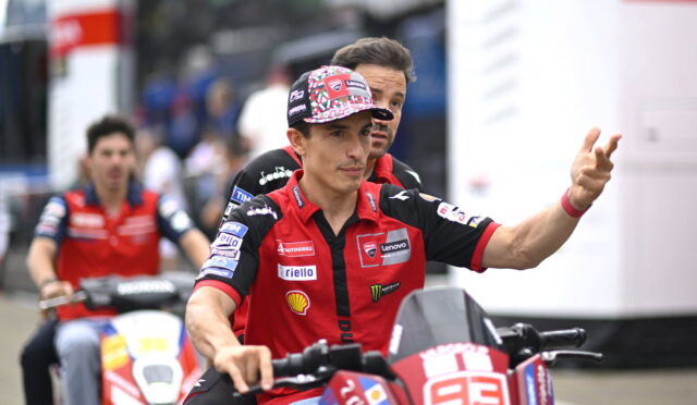 Racing rider in red and black gear on a red bike waves to the crowd, with a teammate on another bike blurred in the background beneath a racing event setting.