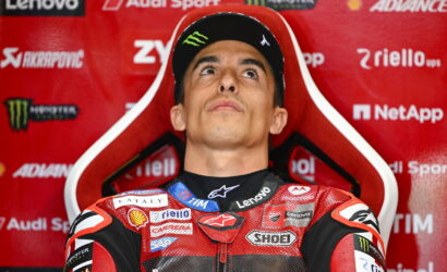 MotoGP rider in a red racing suit sits in a high-backed chair, looking up with a backdrop full of sponsors behind him.