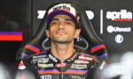 Moto racer wearing a black race suit with purple and red accents, sitting in a racing chair, wearing an Aprilia cap with bold logo.