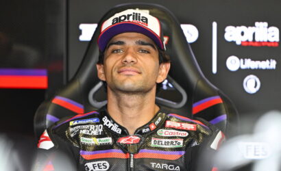Moto racer wearing a black race suit with purple and red accents, sitting in a racing chair, wearing an Aprilia cap with bold logo.