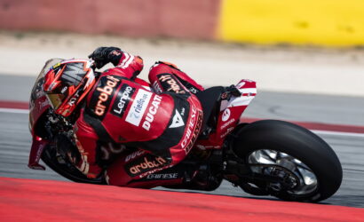 Red Ducati racing motorcycle with rider in red suit leaning into a high-speed corner on a race track, tires gripping the turn.