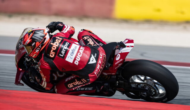 Red Ducati racing motorcycle with rider in red suit leaning into a high-speed corner on a race track, tires gripping the turn.