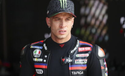 Portrait of a male motorcyclist in a black racing suit with sponsor patches, wearing a leopard-patterned cap with a green Monster logo.