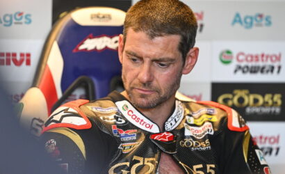 Male motorcycle racer in a black and gold racing suit sits in a paddock, looking down pensively, with sponsor logos like Castrol and GODS45 on his suit and backdrop in view.