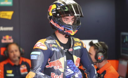 MotoGP rider in a Red Bull helmet and KTM racing suit stands in a team garage with crew members nearby.