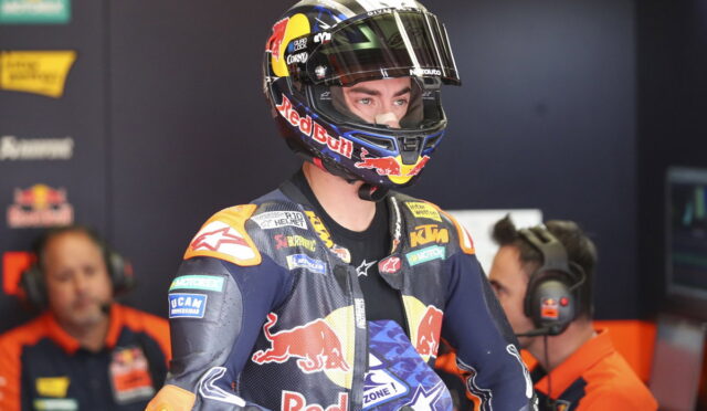 MotoGP rider in a Red Bull helmet and KTM racing suit stands in a team garage with crew members nearby.