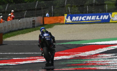 Rear view of a Yamaha racing bike and rider on a track, leaning into a turn with red and white painted curbs and Michelin banners in the background.