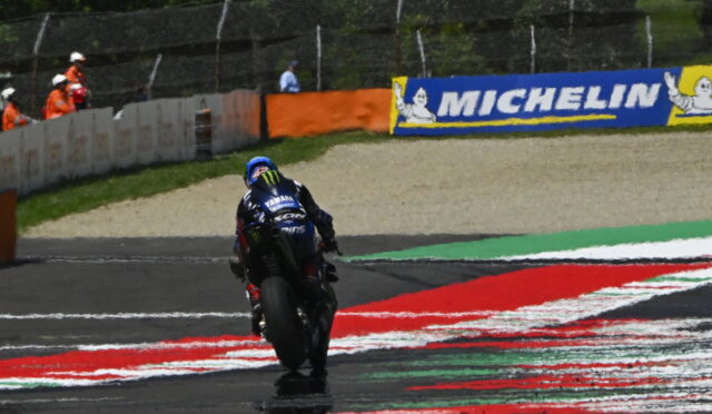 Rear view of a Yamaha racing bike and rider on a track, leaning into a turn with red and white painted curbs and Michelin banners in the background.