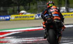 Racing motorcycle rider in orange/black gear leaning into a turn on a track, with Michelin banners along the barriers in the background.