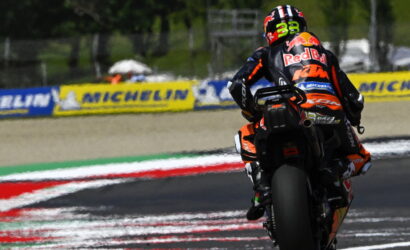 Racing motorcycle rider in orange/black gear leaning into a turn on a track, with Michelin banners along the barriers in the background.