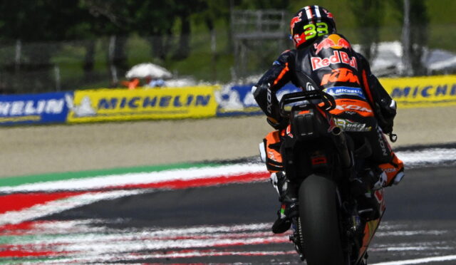 Racing motorcycle rider in orange/black gear leaning into a turn on a track, with Michelin banners along the barriers in the background.