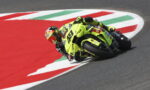 Motorcycle racer in bright yellow gear leans into a sharp turn on a race track.