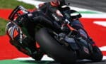 MotoGP rider in red and black leans aggressively into a corner on a high-speed racing bike on a red race track surface with sponsor decals visible on the bike and suit.