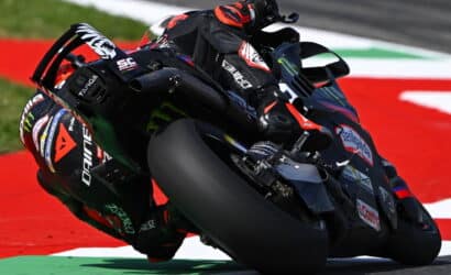 MotoGP rider in red and black leans aggressively into a corner on a high-speed racing bike on a red race track surface with sponsor decals visible on the bike and suit.