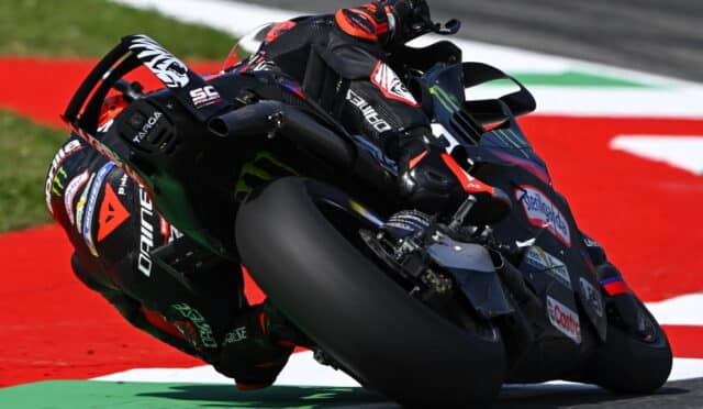 MotoGP rider in red and black leans aggressively into a corner on a high-speed racing bike on a red race track surface with sponsor decals visible on the bike and suit.