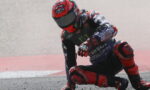 Motorcycle racer in a red helmet crashes and leans on the racetrack beside a second rider in red and blue gear, during a race.