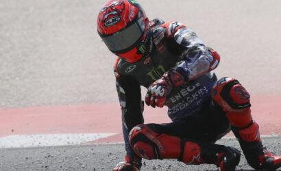 Motorcycle racer in a red helmet crashes and leans on the racetrack beside a second rider in red and blue gear, during a race.