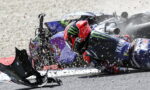 Racer in a red helmet slides along the track after a crash, with debris and a toppled bike nearby.