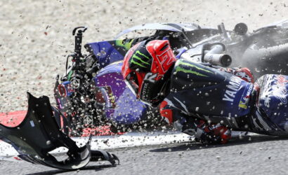 Racer in a red helmet slides along the track after a crash, with debris and a toppled bike nearby.
