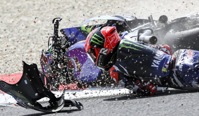 Racer in a red helmet slides along the track after a crash, with debris and a toppled bike nearby.
