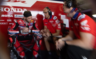 MotoGP rider in red Honda racing suit sits in the pit area with hands clasped while two team members in headsets talk nearby in a red Honda pit wall backdrop.