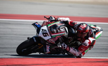 Racing motorcycle rider in red and white gear leans into a sharp turn on a race track; bike shows Italian flag colors and sponsor logos on the fairing.