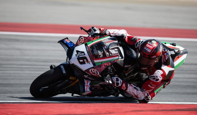 Racing motorcycle rider in red and white gear leans into a sharp turn on a race track; bike shows Italian flag colors and sponsor logos on the fairing.