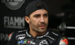 Portrait of a male motorcyclist in a black Dainese racing suit, wearing a backward cap, looking off to the side in a pit area with sponsor logos visible.