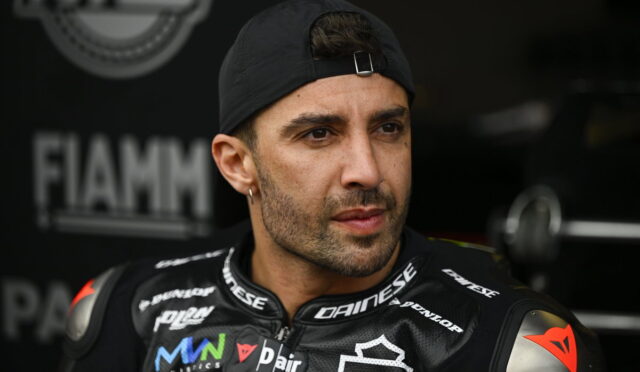 Portrait of a male motorcyclist in a black Dainese racing suit, wearing a backward cap, looking off to the side in a pit area with sponsor logos visible.
