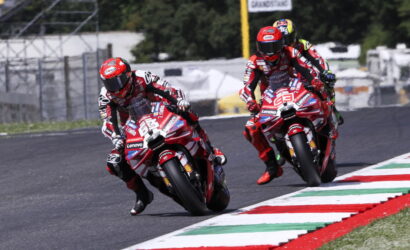 Two motorcycle racers in red gear lean into a curve on a race track, red-and-white bikes covered in sponsor logos.