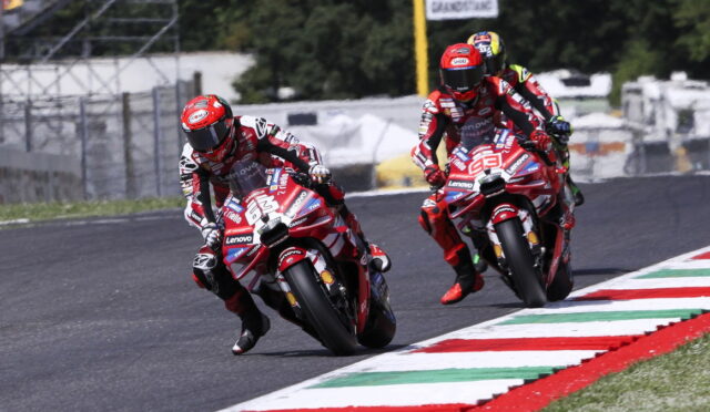 Two motorcycle racers in red gear lean into a curve on a race track, red-and-white bikes covered in sponsor logos.