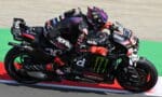 Two MotoGP riders in black Aprilia livery lean into a tight turn on a race track, close together and mid-race.”,