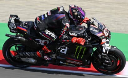 Two MotoGP riders in black Aprilia livery lean into a tight turn on a race track, close together and mid-race.”,