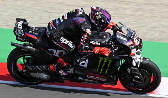 Two MotoGP riders in black Aprilia livery lean into a tight turn on a race track, close together and mid-race.”,